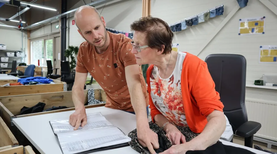 Een vrouw zit rechtsvoor in beeld. Ze zit aan een tafel en kijkt samen met een mannelijke collega naar informatie die in een map staat